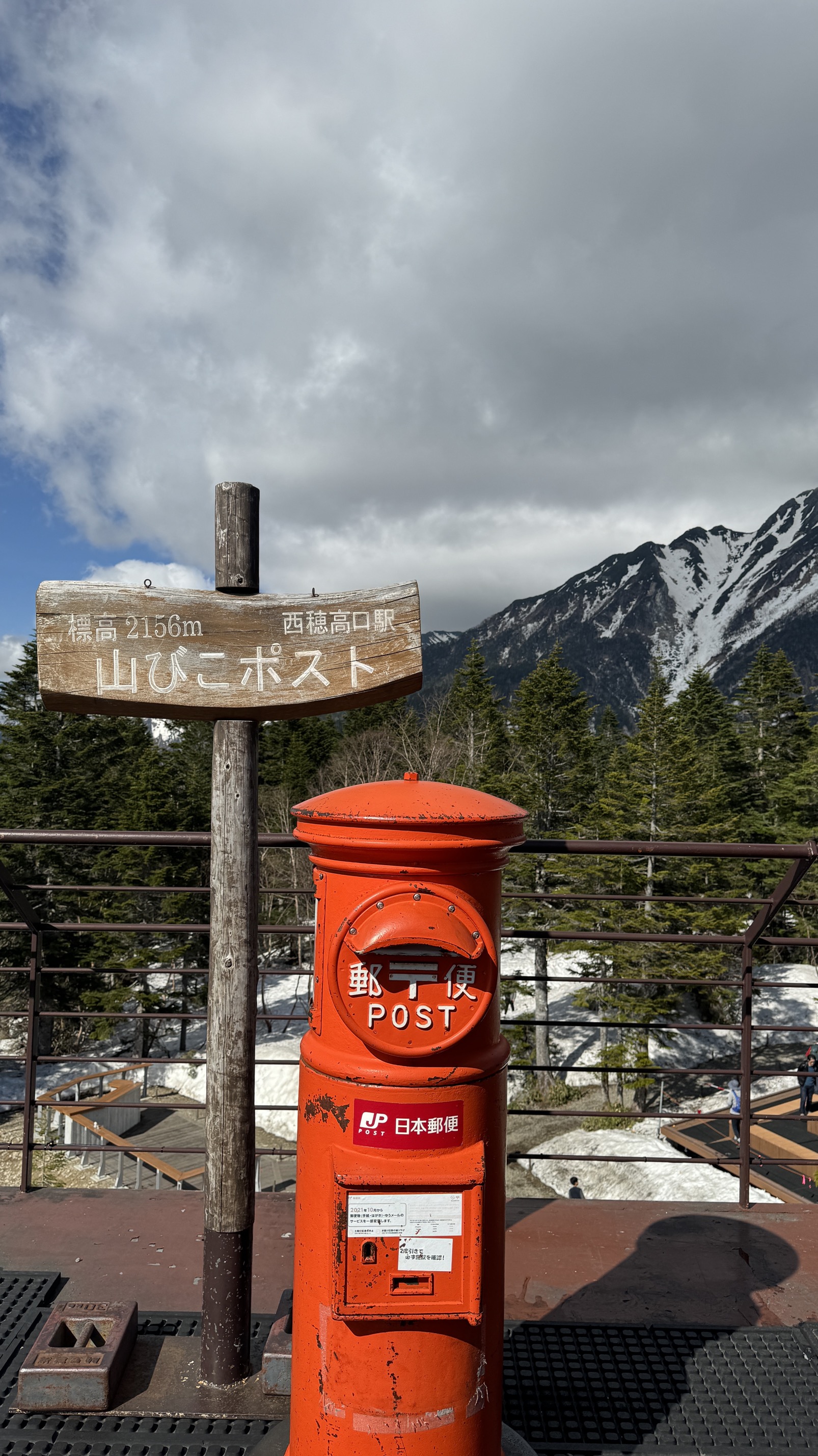 Red postal box at high-altitude trail junction with snow patches and forest, Japanese Alps